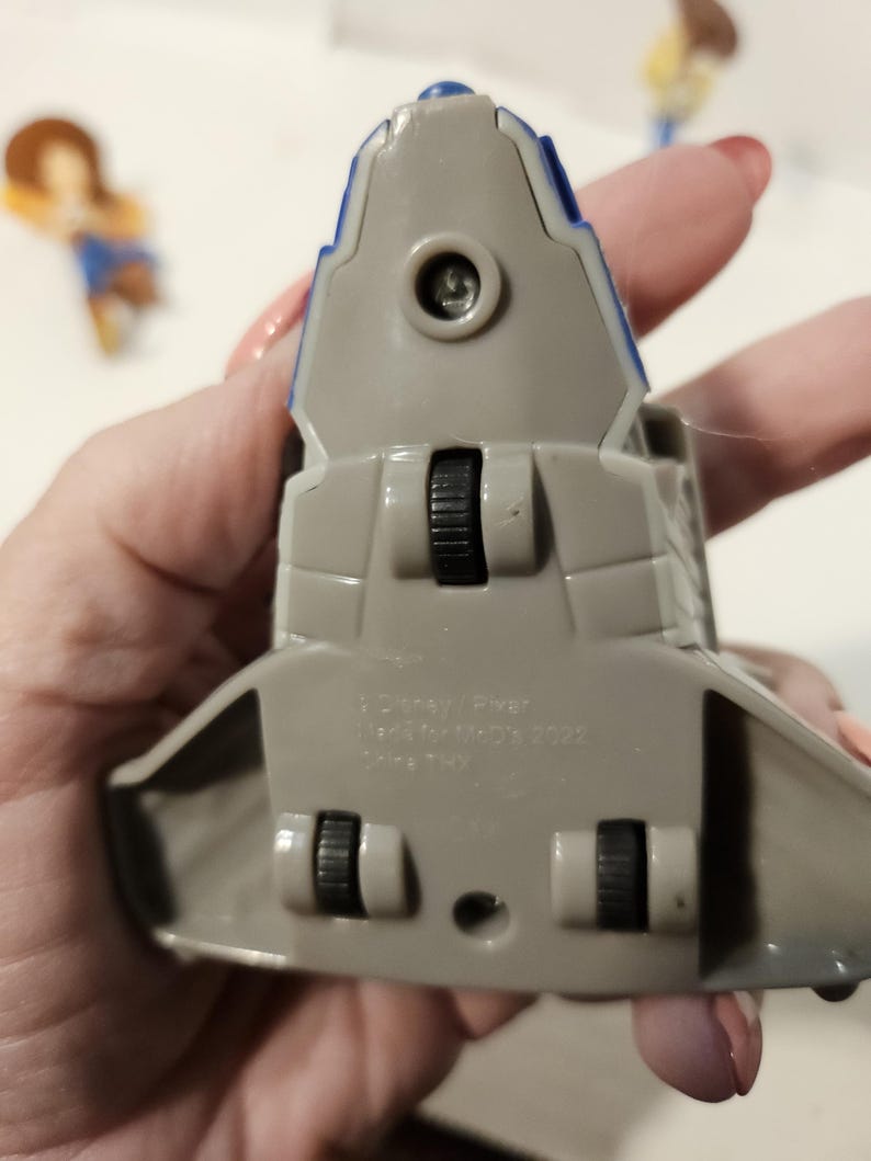 May include: A gray and blue toy spaceship with black wheels. The toy has a blue stripe along the top and is marked with text. The toy is a collectible item.