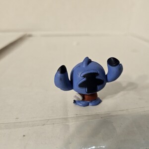Pu&ograve; includere: Piccola statuina blu del personaggio Stitch del film d'animazione Lilo & Stitch. La statuina ha grandi orecchie nere, un segno nero a forma di croce sulla testa e una fascia rossa in vita. La statuina &egrave; su una superficie chiara.