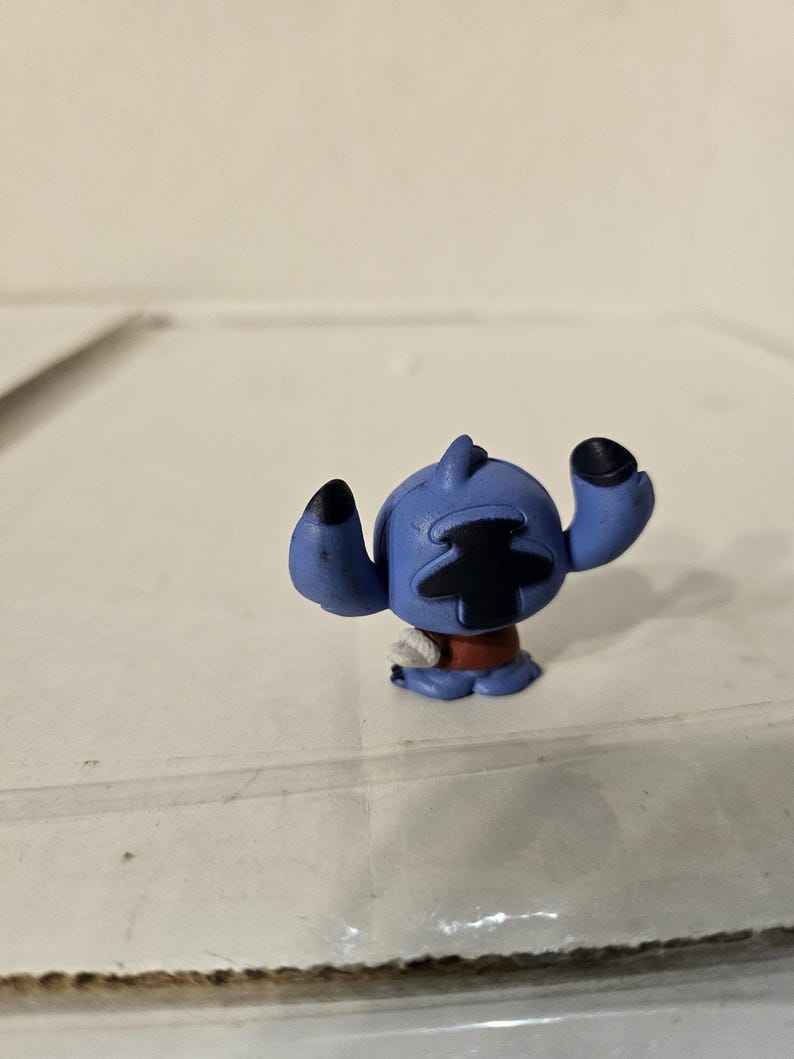Pu&ograve; includere: Piccola statuina blu del personaggio Stitch del film d'animazione Lilo & Stitch. La statuina ha grandi orecchie nere, un segno nero a forma di croce sulla testa e una fascia rossa in vita. La statuina &egrave; su una superficie chiara.