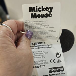 May include: A white tag with the words "Mickey Mouse" in large black letters. The tag includes the Fisher-Price logo and website address. The tag also includes safety information and material details.