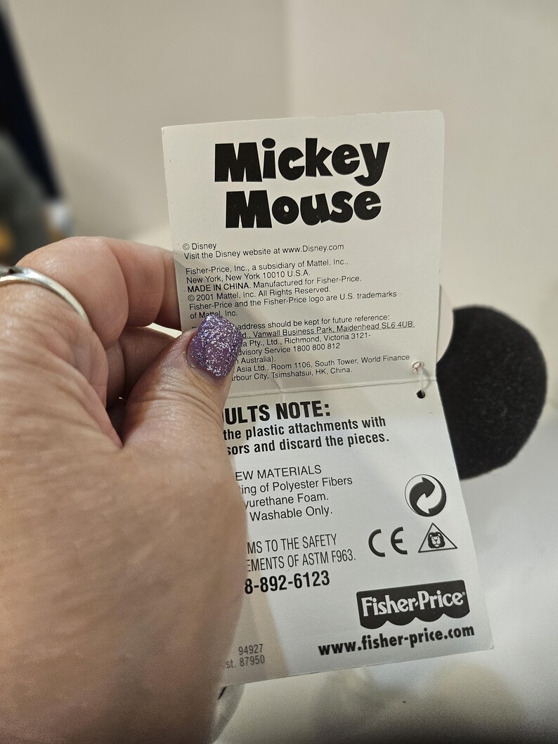 May include: A white tag with the words "Mickey Mouse" in large black letters. The tag includes the Fisher-Price logo and website address. The tag also includes safety information and material details.