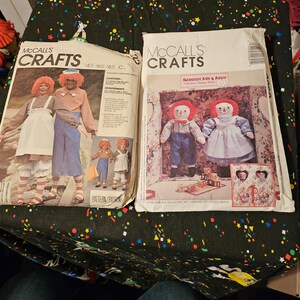 Może przedstawiać: Dwa vintage'owe wzory do szycia McCall's Crafts. Pierwszy wzór to strój Raggedy Ann i Andy. Drugi wzór to lalka Raggedy Ann i Andy. Oba wzory zawierają ilustracje lalek.