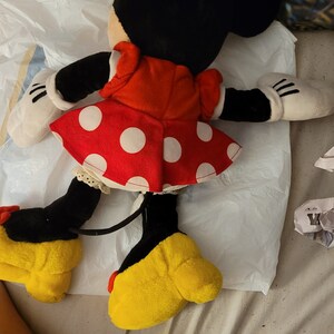 Puede incluir: Un juguete de peluche de Minnie Mouse con un vestido rojo con lunares blancos y zapatos amarillos. El juguete tiene orejas negras y una cola negra.