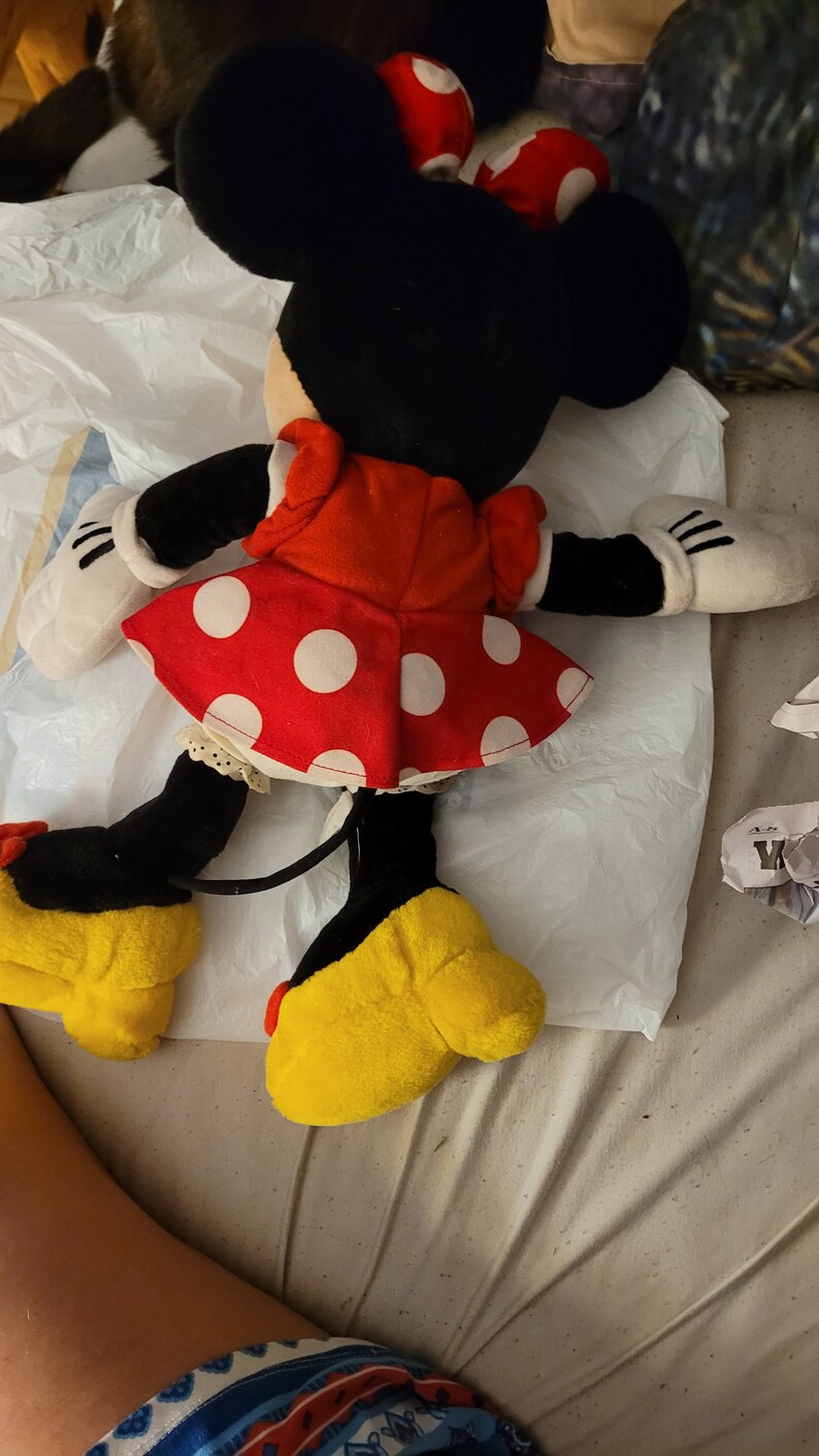 Puede incluir: Un juguete de peluche de Minnie Mouse con un vestido rojo con lunares blancos y zapatos amarillos. El juguete tiene orejas negras y una cola negra.