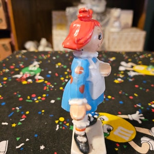 May include: A red-haired doll with a blue dress and white apron. The doll is standing on a white base and has a painted-on smile.