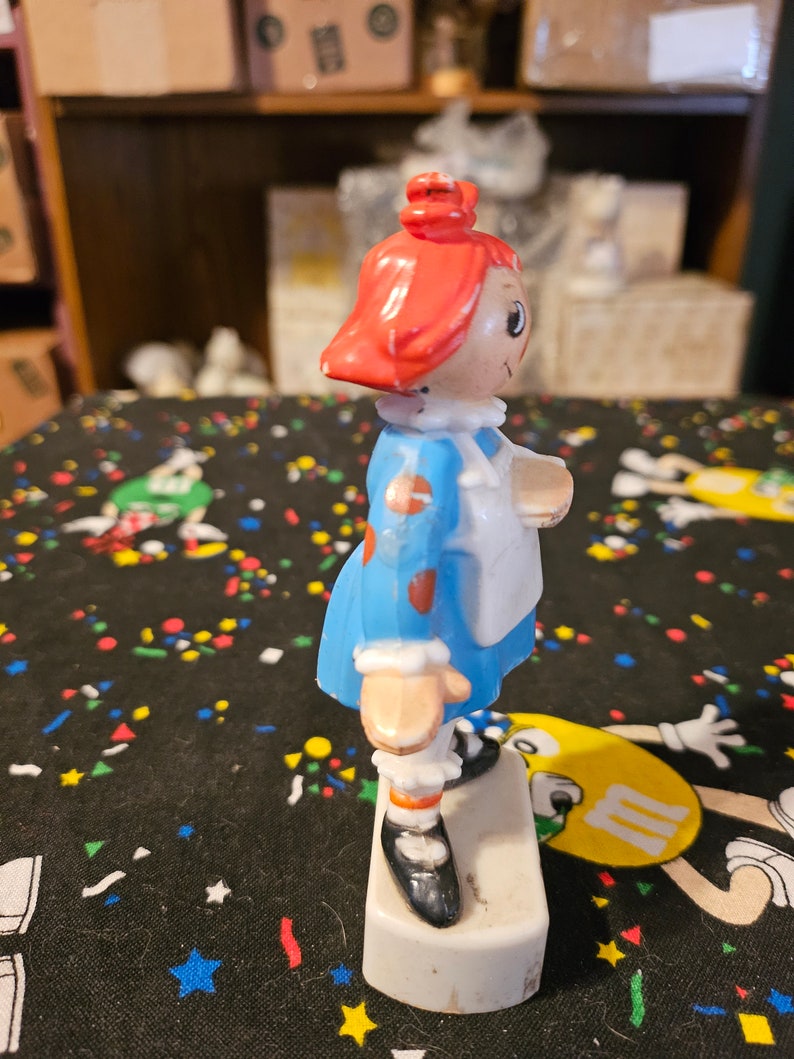 May include: A red-haired doll with a blue dress and white apron. The doll is standing on a white base and has a painted-on smile.