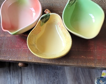 May include: Three small ceramic bowls shaped like fruit. The bowls are pink, green, and yellow and have a glossy finish. The bowls are perfect for serving snacks or dips.
