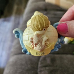 May include: A porcelain figurine of a girl holding a globe with the word "Hope" on a banner. The figurine is painted in shades of white, yellow, blue, and pink.
