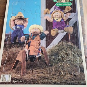 Könnte beinhalten: Ein 25-teiliges Puzzle mit drei Cabbage Patch Kids Puppen. Die Puppen sind in farbenfrohen Outfits gekleidet und stehen in einer Heuballenkulisse. Das Puzzle ist für Kinder von 3 bis 7 Jahren geeignet.