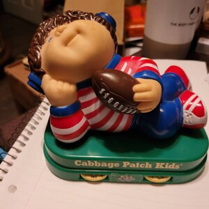 May include: A Cabbage Patch Kids alarm clock featuring a red, white and blue football player holding a brown football. The clock has a green base with three knobs labelled "160", "320", and "Off".