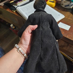 K&ouml;nnte beinhalten: Eine schwarze Samt-Puppe mit schwarzen Haaren, die einen langen schwarzen Mantel tr&auml;gt. Die Puppe wird von einer Hand gehalten, die ein Armband tr&auml;gt.
