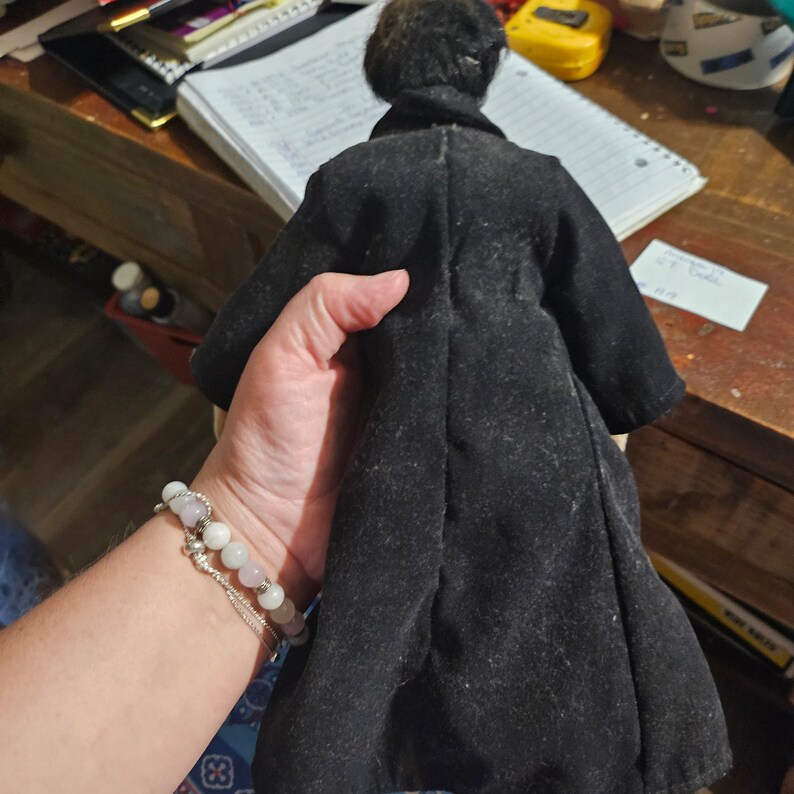 K&ouml;nnte beinhalten: Eine schwarze Samt-Puppe mit schwarzen Haaren, die einen langen schwarzen Mantel tr&auml;gt. Die Puppe wird von einer Hand gehalten, die ein Armband tr&auml;gt.