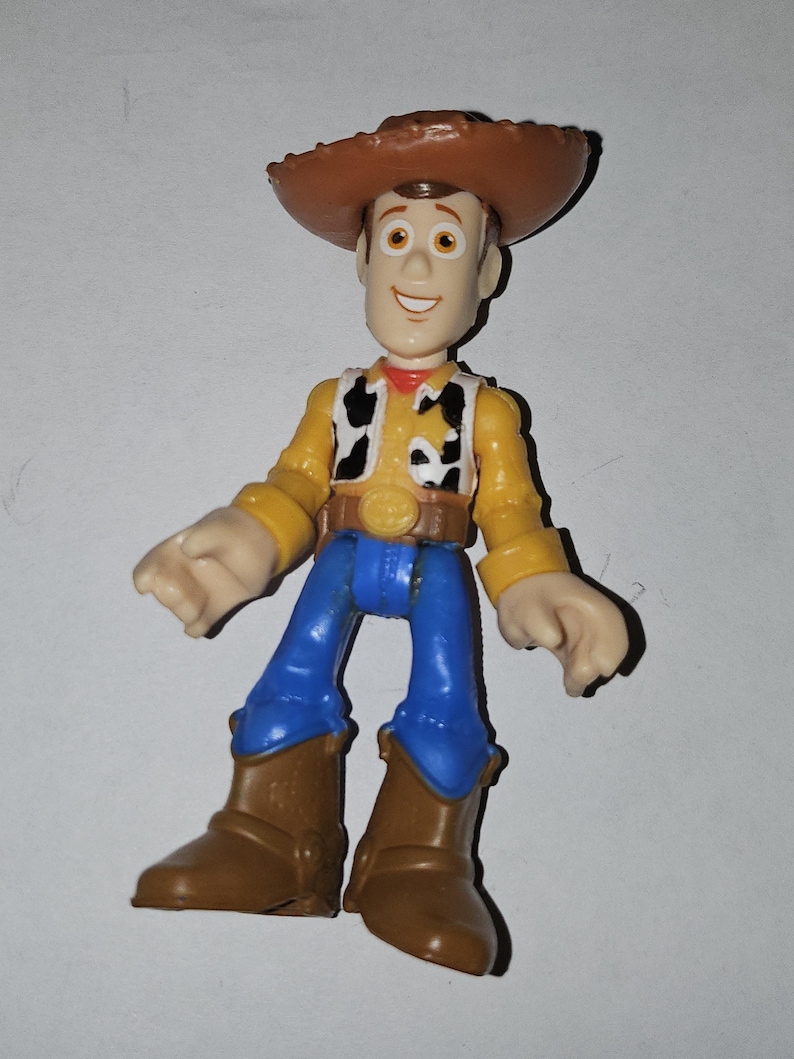 Op de afbeelding: Een plastic speelgoedfiguur van een cowboy, met een bruine hoed, geel overhemd, zwart-wit vest, blauwe broek en bruine laarzen. De figuur heeft een lachend gezicht. Een verzamelobject.