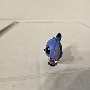 Pu&ograve; includere: Piccola statuina blu del personaggio Stitch del film d'animazione Lilo & Stitch. La statuina ha grandi occhi neri, grandi orecchie e un abito marrone. La statuina &egrave; in piedi su una superficie trasparente.