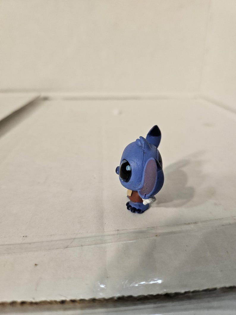 Pu&ograve; includere: Piccola statuina blu del personaggio Stitch del film d'animazione Lilo & Stitch. La statuina ha grandi occhi neri, grandi orecchie e un abito marrone. La statuina &egrave; in piedi su una superficie trasparente.