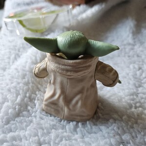 Puede incluir: Una peque&ntilde;a figura de piel verde con grandes orejas, vestida con una t&uacute;nica color beige. La figura es un personaje de una popular franquicia de ciencia ficci&oacute;n. La figura est&aacute; colocada de espaldas al espectador, sobre una superficie blanca y texturizada.