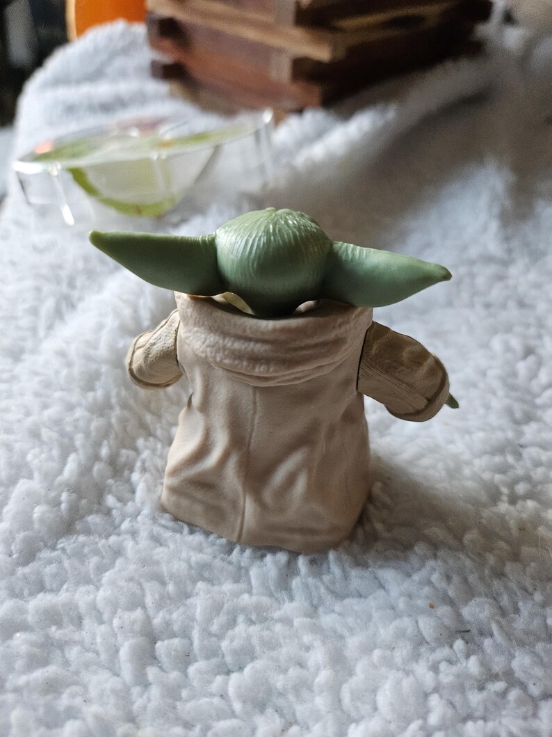 Puede incluir: Una peque&ntilde;a figura de piel verde con grandes orejas, vestida con una t&uacute;nica color beige. La figura es un personaje de una popular franquicia de ciencia ficci&oacute;n. La figura est&aacute; colocada de espaldas al espectador, sobre una superficie blanca y texturizada.