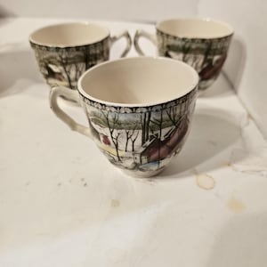 May include: Three white ceramic teacups with a winter scene design. The cups have a brown, green, and red color scheme and feature a snowy landscape with houses and trees.