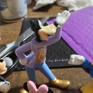 Może przedstawiać: Figurka Goofy'ego w fioletowej kurtce ze złotymi akcentami, niebieskich spodniach i złotych butach. Jego prawa ręka jest uniesiona, ma czarną głowę i długi, brązowy pysk. Częściowo widoczne są inne figurki postaci z kresk&oacute;wek.
