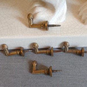 May include: Six antique brass L-shaped furniture pulls with decorative finials and attached screws. The pulls are arranged on a grey surface, with a white ceramic object in the background. These vintage hardware pieces are likely for cabinets or drawers.