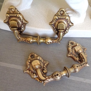 May include: Three antique brass drawer pulls with ornate floral designs. Each pull features a curved handle and decorative backplate. The brass has a warm, aged patina, suggesting vintage craftsmanship. These pulls are ideal for adding a touch of vintage charm to furniture.