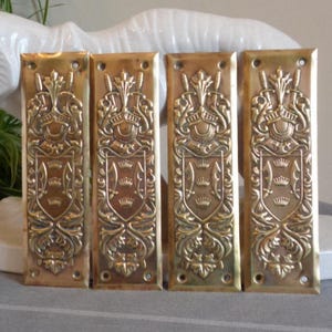 Può includere: Quattro placche per porte antiche in ottone con disegni in rilievo. Ogni placca presenta uno scudo con tre corone, un elmo e spade incrociate. Le placche sono rettangolari con bordi arrotondati e fori preforati.