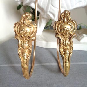 May include: A pair of gold-toned metal curtain tiebacks featuring ornate, symmetrical scrollwork and leaf-like detailing at the top. Each tieback has a curved, slender arm extending downwards from the decorative head, finished in a metallic gold.