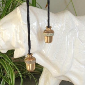 Puede incluir: Dos pequeñas cadenas doradas con cordones negros cuelgan frente a una escultura de cerámica blanca. Las cadenas tienen una banda decorativa y texturizada en el medio. La escultura es una forma animal estilizada.