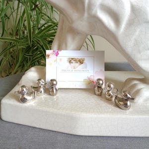 Peut inclure: Plusieurs figurines de canards argentés et des figures abstraites sont disposées sur une surface en céramique blanche craquelée. Une carte avec le texte "FRENCH MEMORIES" et un portrait est également présente. Une sculpture blanche est en arrière-plan.