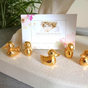 Peut inclure: Plusieurs figurines de canards dorés disposées sur un plateau blanc à surface craquelée. Une carte avec le texte "FRENCH MEMORIES" et un portrait vintage est en arrière-plan. Les canards sont dans différentes poses.
