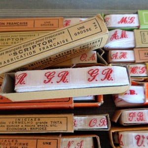 Può includere: Una collezione di scatole di cartone vintage contenenti nastro di cotone bianco con iniziali ricamate in rosso. Le scatole presentano testo in francese tra cui "SCRIPTOR" e "FABRICATION FRANÇAISE", con una scatola aperta che mostra il monogramma rosso "G.R.".