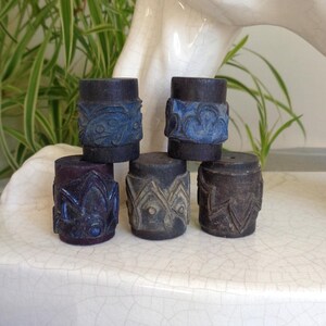 May include: A collection of five cylindrical wooden stamps, each featuring distinct carved patterns. Two stamps display blue-tinted floral motifs, while the remaining three show geometric and leaf-like carvings in a dark, natural wood finish.
