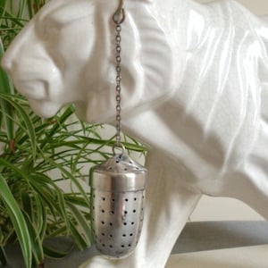 May include: A silver-toned metal tea infuser with a perforated acorn-shaped body and a small lid, suspended by a thin metal chain from a hook. The infuser features a series of small circular holes across its surface for steeping loose leaf tea.