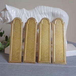 May include: Four rectangular brass door plates with a textured gold finish and a rounded top arch. Each plate features a decorative beaded border along the edges, providing a uniform appearance for these architectural hardware components.