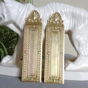 Puede incluir: Dos placas de puerta rectangulares de latón con un borde en relieve adornado con un patrón de hojas y un elemento decorativo en forma de abanico en la parte superior. Cada placa tiene un panel central liso y orificios para montaje.