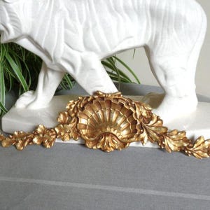 Könnte beinhalten: Ein goldfarbenes dekoratives Wandornament mit einem zentralen Jakobsmuschel-Motiv, das von symmetrischen Akanthusblatt-Schnitzereien flankiert wird. Das Stück hat eine metallische Oberfläche mit detaillierten Reliefverzierungen.
