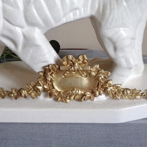 May include: A gold-toned metal decorative plaque featuring an ornate floral garland design with a central bow detail. The piece is positioned in front of the white ceramic legs of a large animal figurine on a flat white base.