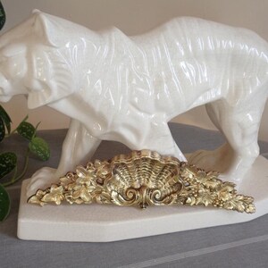 May include: A white ceramic tiger figurine standing on a matching base, featuring a glossy finish and molded stripes. Positioned in front of the tiger is an ornate, gold-colored metal decorative piece shaped like a shell with intricate leaf details.