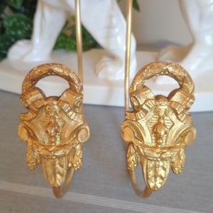 Peut inclure: Une paire d'embrasses à rideaux en métal doré présentant une boucle supérieure décorative et un motif central en relief avec des volutes symétriques et des éléments végétaux. Chaque embrasse possède un bras métallique incurvé.