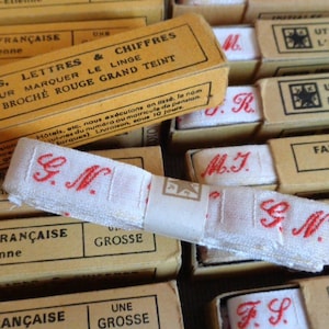 Könnte beinhalten: Nahaufnahme von Vintage-Leineninitialen und -zahlen zum Markieren von Stoff. Das weiße Stoffband ist mit roten Initialen bestickt und in kleinen, hellbraunen Schachteln verpackt. Die Schachteln haben französischen Text.