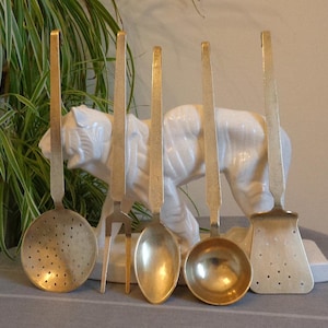 May include: A collection of five brass kitchen tools: a slotted spoon, fork, spoon, ladle, and slotted spatula. They are displayed in front of a white ceramic tiger statue and a green leafy plant.