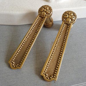 Può includere: Due maniglie antiche in ottone. Ogni maniglia presenta un corpo rettangolare con una superficie strutturata e una fila di piccole perle rotonde lungo i bordi. La parte superiore di ogni maniglia presenta un design circolare con dettagli floreali. Le maniglie sono su uno sfondo azzurro.