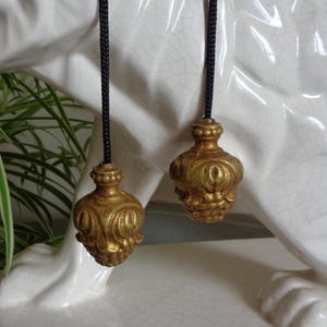 Può includere: Primo piano di due catene di trazione dorate e ornate con terminali decorativi. I terminali hanno un design dettagliato e strutturato e sono sospesi da cordoncini neri. Lo sfondo è una superficie di ceramica bianca screpolata.