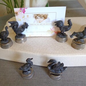 May include: A selection of antique metal bottle stoppers with bird designs, including roosters, hens, ducks, and swans. The stoppers have a silver base and are displayed on a white surface. A card with the text "FRENCH MEMORIES" is visible in the background.