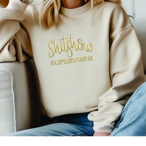May include: A beige sweatshirt with the text "Shit Show Supervisor" in gold lettering.