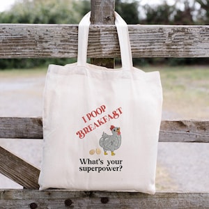 May include: Off-white canvas tote bag with the text "I POOP BREAKFAST" in red script. Below the text is a cartoon chicken with two eggs. The text "What's your superpower?" is below the chicken.
