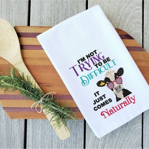 May include: White kitchen towel with a black and white cow wearing pink sunglasses. The text on the towel reads "I'm not trying to be difficult, it just comes naturally."
