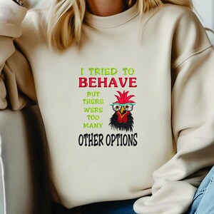Puede incluir: Sudadera beige con el texto "Intenté portarme bien, pero había demasiadas otras opciones". El diseño presenta un pollo de dibujos animados con gafas. El texto es en verde, rojo y negro.