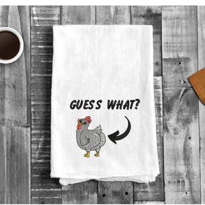 May include: White kitchen towel with a gray embroidered chicken and the text "GUESS WHAT?" with a black arrow pointing to the chicken's rear end.
