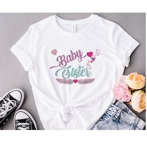 May include: White t-shirt with the words "Baby Sister" in a script font, accented with pink and teal details. The design includes hearts and feather-like elements. The shirt is paired with denim shorts and black sneakers.